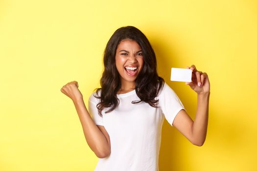 Portrait of cheerful african-american female model, showing credit card and shouting for joy, happy about shopping, standing over yellow background.