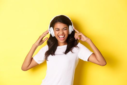 Portrait of modern african-american girl, enjoying listening to music in headphones, smiling amazed, standing over yellow background.