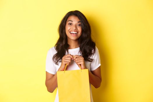 Portrait of cheerful african-american girl shopping, opening a bag with gift and smiling happy, standing over yellow background.