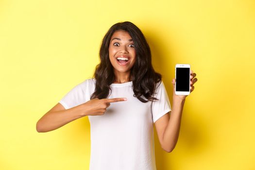 Image of beautiful african-american girl, smiling and looking excited while pointing at smartphone screen, standing over yellow background.