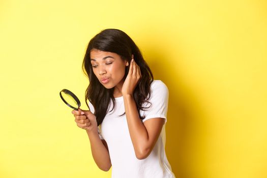 Image of cute african-american woman searching for something with magnifying glass, looking down, standing over yellow background.
