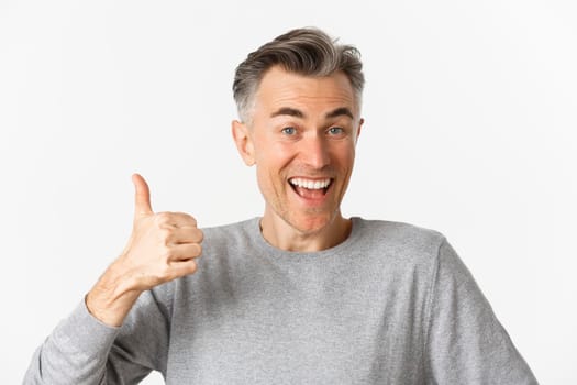 Close-up of cheerful middle-aged man, smiling happy and satisfied, showing thumbs-up, expressing approval, like something good, standing over white background.