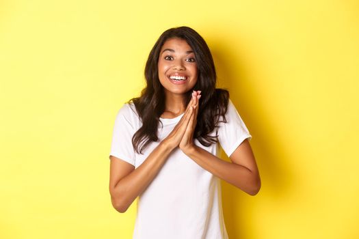 Image of happy and pleased african-american girl, clap hands and looking excited, thanking for something, standing over yellow background.