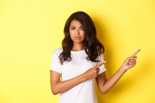 Portrait of skeptical and unamused african-american girl, judging something bad, pointing fingers right with disappointed expression, standing over yellow background.