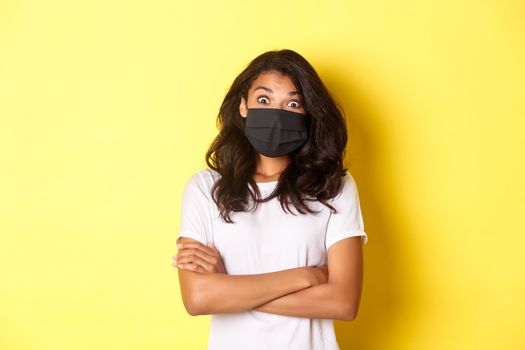 Concept of coronavirus, pandemic and lifestyle. Image of surprised african-american girl in face mask, looking amazed at something cool, standing over yellow background.