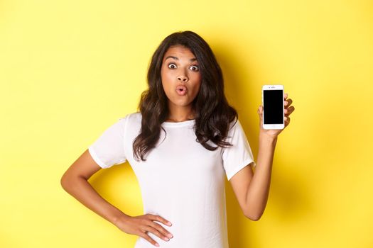 Image of amazed african-american girl, looking fascinated and showing smartphone screen, standing over yellow background.