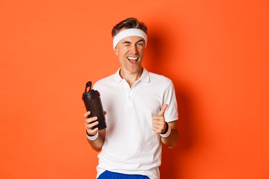 Portrait of confident, handsome middle-aged man in workout uniform, drinking water, winking and showing thumbs-up in approval, standing over orange background.