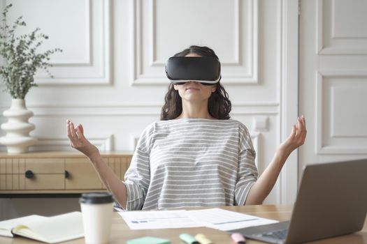 Calm peaceful woman employee meditating in glasses of virtual reality, sitting in zen pose at workplace with laptop computer while working in office, mindful female practicing yoga in VR headset