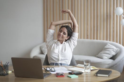 Tired positive italian woman remote employee stretching arms while sitting at desk with laptop and working on deadline from home office. Busy female freelancer doing overtime job