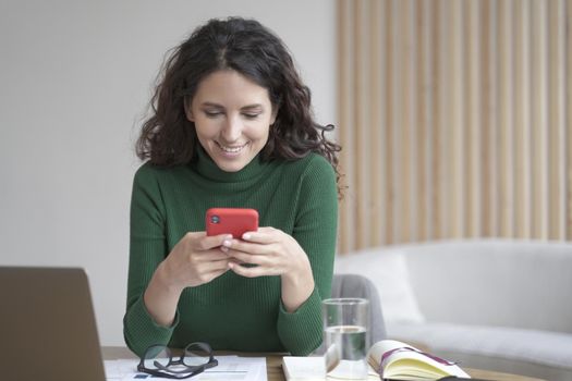 Young beautiful italian freelancer lady with dark wavy hair holding smartphone and looking at screen with smile, texting while sitting with laptop, account papers and bills at modern workplace at home