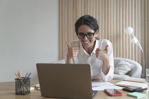 Confident business lady looking at `computer screen with broad smile while showing thumbs-up with both hands. Spanish female tutor working on laptop at home office. Remote work concept