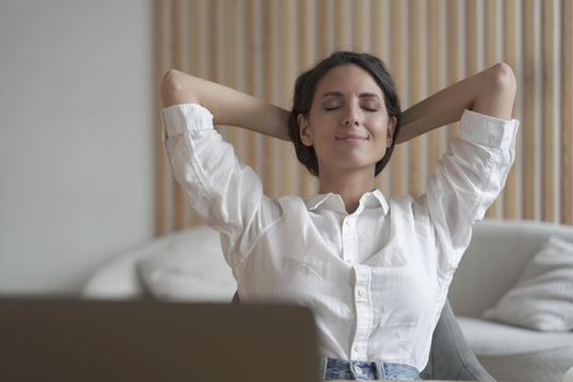 Attractive positive young female entrepreneur sitting at desk at home in relaxing position with hands behind her head and eyes closed, resting between training sessions while working remotely online