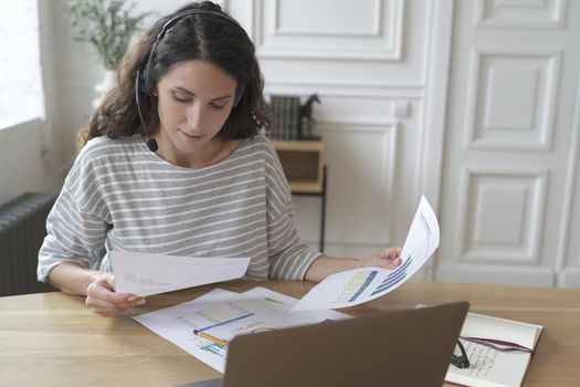 Focused european woman accountant in headset checking documents, charts and reports while using laptop at home office. Concentrated lady preparing financial report for meeting, analyzing statistics