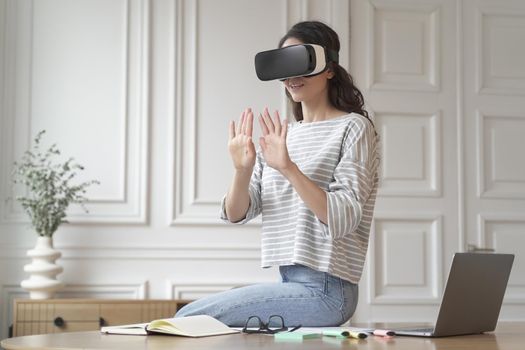 Young excited woman freelancer sitting on top of desk wearing VR glasses, hands raised in air seemingly against invisible wall in 3D game while interacting with digital interface in virtual reality