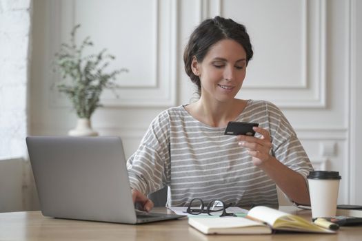 Online shopping. Young smiling italian woman in casual clothes looking at credit card and typing on laptop, paying online, buying goods or services in internet store while sitting at table at home