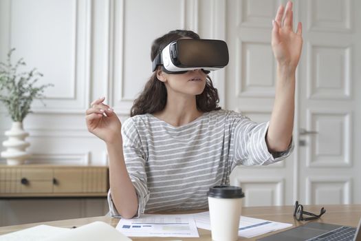 Young woman bank accountant in VR headset sitting at desk at home office, interacting with digital interface while testing and using innovative device for business. Cyberspace experience concept