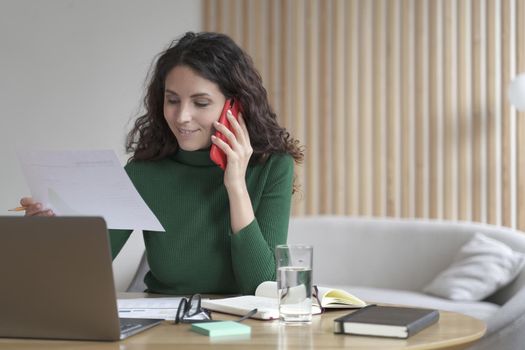 Young smiling spanish businesswoman reading paper document and talking via mobile phone with client while working remotely at home, pleased female calling business partner to discuss sales report