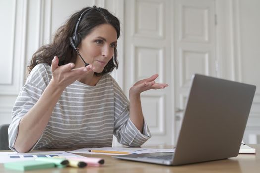 Young smiling Italian woman freelancer in headset gesturing while consulting client customer via video call on laptop, pleasant mixed-race female tutor giving online lesson talking with students
