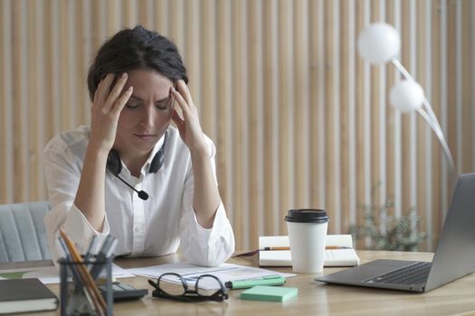 Tired spanish businesswoman with closed eyes sitting at workplace in office and suffering from headache, upset female employee holding head in hands and feeling sad about financial problems at work