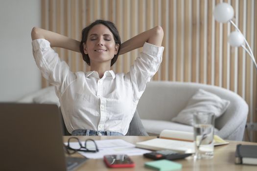 Positive Spanish woman rests during remote work online at home. Freelance lady sitting in front of laptop in relaxed position with satisfied smile on her face while eyes closed and hands behind head