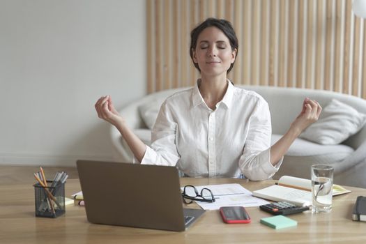 Calm italian female sits in relaxed yoga position at desk in front of laptop with eyes closed while folding her fingers in mudra for better concentration and memory during remote work at home