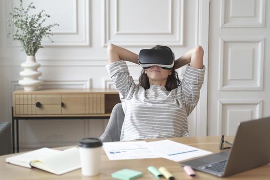 Young glad relaxed woman office worker in virtual reality glasses at workplace with laptop computer, smiling impressed female employee relaxing in VR helmet during break at work