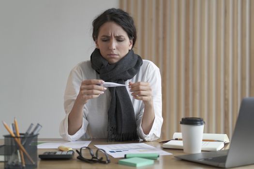 Sick unhealthy Italian woman office worker looking at thermometer, suffer of flu influenza while sitting at her workplace. Ill female employee measuring body temperature at work