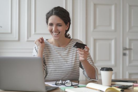 Young happy Spanish woman holding credit card and feeling excited while doing online shopping on laptop at home, looking at computer screen and purchasing goods or services in internet store