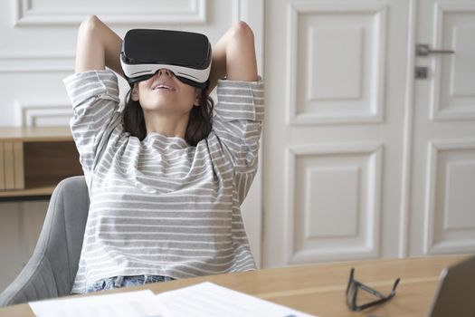 Young happy female employee in VR goggles at workplace watching movies, relaxed woman office worker sitting at desk wearing virtual reality helmet and enjoying 3D experience at work