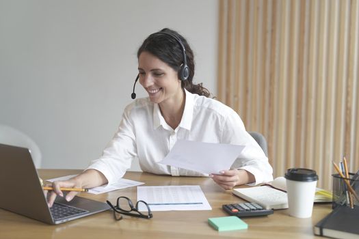 Happy millennial woman wears headphones while sitting at desk at home office looks at laptop screen works online. Smiling young female tutor typing on computer, customer couching distant on web