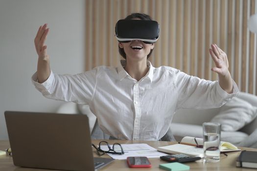 Excited european business woman testing VR glasses at work, sitting at her workplace with laptop in office and using innovative virtual reality technology, female worker playing 3d game during break