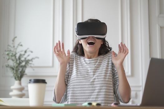 Excited cheerful entrepreneur lady testing virtual reality technology wearing VR glasses sitting at home office, interacting with 360 degrees headset while raised hands up and opened mouth from joy
