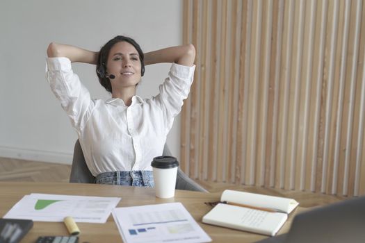 Meditative contented female entrepreneur in wireless headset sitting in relaxed position with hands behind head, looking sideways, taking break between customer calls, while working remotely online