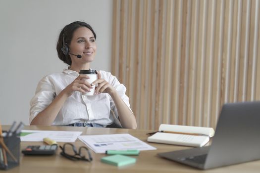 Pleasant attractive young Italian businesswoman in wireless headset sitting at desk at home office leaning back on chair with takeaway coffee in hands enjoying hot drink while working remotely online