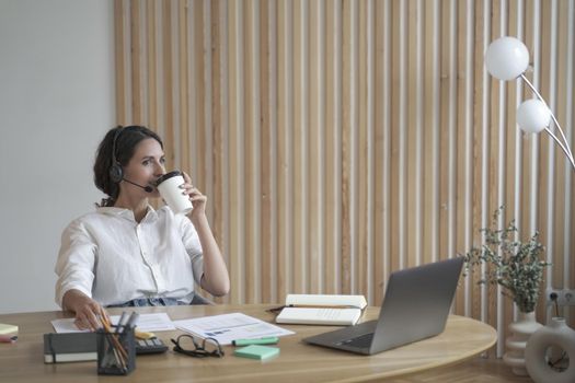 Satisfied positive Spanish female entrepreneur in wireless headphones drinking coffee from disposable cup while sitting in front of laptop at modern homeplace, leaning back and enjoying moment of rest