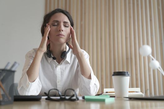 Frustrated Italian woman with closed eyes trying to concentrate while sitting at her workplace in modern office, tired Spanish female employee massaging temples, suffering from headache after work