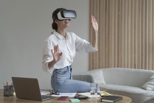 Photo of businesswoman in elegant formal clothes sitting in office and using VR technologies at work, female employee wearing 3d goggles engaged in virtual conference with colleagues