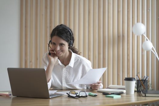 Happy young hispanic business woman in headset with microphone speaking by video call, looking at laptop computer while sitting at her workplace in modern office, call center agent consulting client