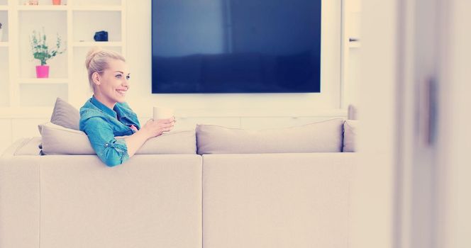 beautiful blonde woman enjoying a cup of coffee while sitting on a sofa in her apartment