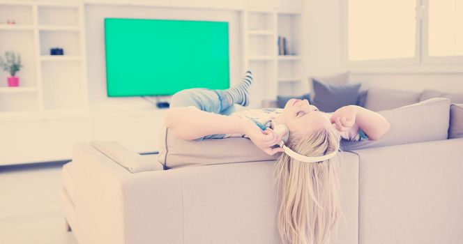 Young beautiful girl enjoying music through headphones, laying on sofa at home