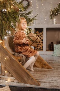 A little blonde girl is sitting on a wooden staircase in a Scandinavian interior decorated in a New Year's style. A child holds a Christmas gift in a craft package.