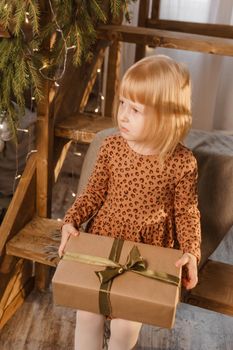 A little blonde girl is sitting on a wooden staircase in a Scandinavian interior decorated in a New Year's style. A child holds a Christmas gift in a craft package.