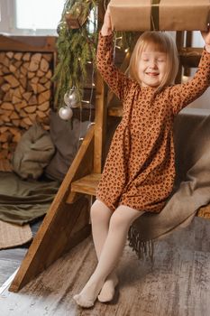 A little blonde girl is sitting on a wooden staircase in a Scandinavian interior decorated in a New Year's style. A child holds a Christmas gift in a craft package.