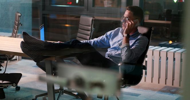 Full length of a relaxed casual young businessman sitting with legs on desk at night office