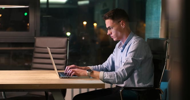 Young man working on laptop at night in dark office. The designer works in the later time.