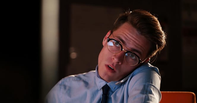 Young man using mobile phone while working on computer at night in dark office.