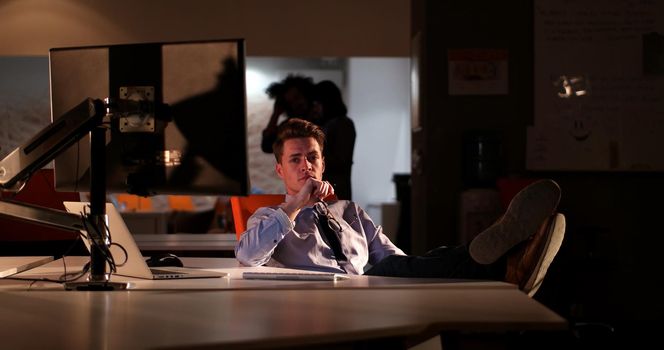 Full length of a relaxed casual young businessman sitting with legs on desk at night office