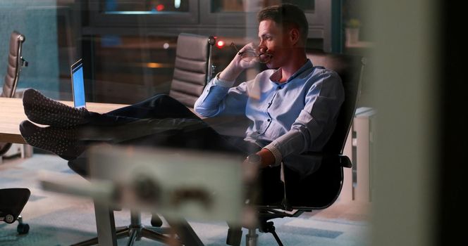 Full length of a relaxed casual young businessman sitting with legs on desk at night office