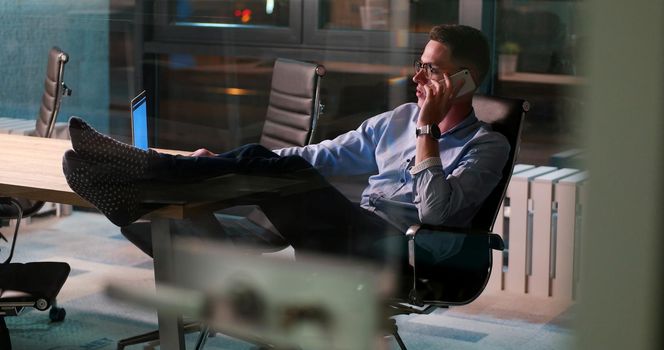 Full length of a relaxed casual young businessman sitting with legs on desk at night office