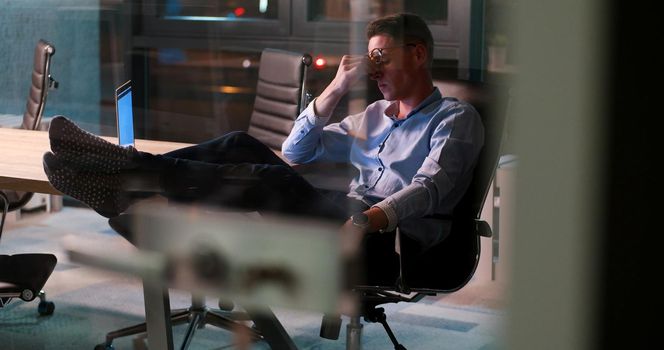 Full length of a relaxed casual young businessman sitting with legs on desk at night office